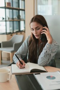 Businesswoman multitasking at office desk, writing notes while speaking on phone.